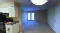 This single-floor space features an open-concept layout that connects the living area to the kitchen. The living room is finished with plush wall-to-wall carpeting in a neutral tone, creating a comfortable and inviting atmosphere. A pair of white-framed French doors dominates the far wall, allowing natural light to filter into the space and providing access to the exterior. The walls are painted in a warm taupe hue, complemented by clean white baseboards that run throughout the room. An overhead light fixture provides additional illumination to the living area. Adjacent to the living space, the kitchen features white cabinetry with a clean, traditional style and dark-toned countertops that offer a subtle contrast. The transition between the kitchen and living area is defined by a structural column, which adds architectural character to the open layout. The overall ambiance of the space is straightforward and functional, with neutral finishes that provide a versatile backdrop suitable for a variety of interior styles and furnishing choices.
