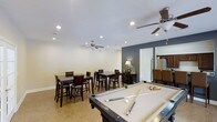 This single-floor space is designed as a versatile entertainment and recreation area, offering a well-appointed layout suited for socializing and leisure. The focal point of the room is a full-sized pool table positioned prominently in the foreground, providing an immediate invitation for recreation. Surrounding it, multiple high-top dining tables paired with dark wood bar-height chairs are arranged throughout the space, accommodating groups comfortably for casual dining or conversation.

The room features neutral-toned walls accented by a striking dark gray feature wall, adding depth and visual contrast to the overall aesthetic. Recessed lighting is installed throughout the ceiling, complemented by two ceiling fans that ensure consistent airflow and comfort across the generous open floor plan. Tile flooring in a warm, neutral hue runs throughout, offering both durability and a clean, polished appearance.

A decorative lamp atop a dark wood console table adds a warm ambient touch to one corner of the room. Adjacent to the main recreation area, a glimpse into what appears to be a kitchen reveals stainless steel appliances and dark cabinetry, extending the functional appeal of this entertainment-focused space.