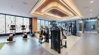 Located at 300 K St in Washington, DC, this expansive fitness center offers a thoughtfully designed, multi-zone exercise facility spread across a single floor. The main exercise room is a standout space, distinguished by a striking coffered ceiling with elegant curved LED lighting that casts a warm, ambient glow throughout. Floor-to-ceiling windows allow ample natural light to flood in while providing views to the outside. The room is fully equipped with a wide array of cardio machines — including treadmills, ellipticals, and stair climbers — as well as selectorized strength machines, cable systems, free weights, benches, and TRX suspension trainers. Vibrant, colorful geometric wall murals add an energetic and modern aesthetic throughout. A secondary exercise room features a dedicated cycling and rowing studio outfitted with Echelon connected fitness equipment, light wood flooring, and subtly textured grey walls. An additional multipurpose studio offers a mirrored barre wall, light hardwood floors, and large windows with decorative typographic glass treatments, making it ideal for yoga, barre, or stretching. A functional turf training zone with plyo boxes, kettlebells, and a BOSU ball rounds out the facility. Together, these spaces create a comprehensive, design-forward fitness environment suited for a wide range of workout styles.