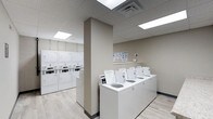 The Laundry Center is a thoughtfully designed, single-floor communal facility built for practicality and ease of use. The space features a clean, neutral color palette with warm beige walls and light wood-look flooring that contribute to a bright, welcoming atmosphere. Recessed panel lighting in the drop-tile ceiling ensures the entire room is evenly and efficiently illuminated throughout.

The facility is equipped with a row of top-loading washing machines neatly arranged along a central counter-height platform, making loading and unloading straightforward. Along the opposite wall, a bank of stacked dryer units maximizes vertical space, offering substantial drying capacity without overcrowding the floor area. Instructional signage is posted on the wall above the washers, providing clear guidance for users.

A structural column divides the space into two functional zones, helping to organize traffic flow between the washing and drying areas. The overall layout is open and uncluttered, allowing for comfortable movement throughout. The combination of modern equipment, clean finishes, and practical design makes this laundry center a highly functional shared amenity.