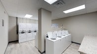 The Laundry Center is a thoughtfully designed, single-floor communal facility built for practicality and ease of use. The space features a clean, neutral color palette with warm beige walls and light wood-look flooring that contribute to a bright, welcoming atmosphere. Recessed panel lighting in the drop-tile ceiling ensures the entire room is evenly and efficiently illuminated throughout.

The facility is equipped with a row of top-loading washing machines neatly arranged along a central counter-height platform, making loading and unloading straightforward. Along the opposite wall, a bank of stacked dryer units maximizes vertical space, offering substantial drying capacity without overcrowding the floor area. Instructional signage is posted on the wall above the washers, providing clear guidance for users.

A structural column divides the space into two functional zones, helping to organize traffic flow between the washing and drying areas. The overall layout is open and uncluttered, allowing for comfortable movement throughout. The combination of modern equipment, clean finishes, and practical design makes this laundry center a highly functional shared amenity.