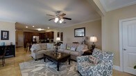 This two-story home showcases a thoughtfully designed open-concept layout, where the living area and kitchen connect to create a cohesive, functional space. The living room is anchored by a large, decorative area rug featuring a bold floral pattern, complementing the neutral tones of the tufted sofa and coordinating accent chairs adorned with patterned throw pillows. A dark wood coffee table serves as a central focal point, adding contrast against the lighter furnishings. Crown molding lines the ceiling, adding an elegant architectural detail, while a ceiling fan with integrated lighting provides both comfort and illumination. Recessed lighting throughout ensures the space remains bright and welcoming. The open kitchen, visible from the living area, features rich dark wood cabinetry, stainless steel appliances, and a breakfast bar, making it ideal for entertaining. A framed mirror on the living room wall reflects light and enhances the sense of openness. The overall ambiance is warm and traditional, with carefully chosen décor that creates a comfortable, family-friendly environment across both floors.