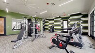 Located at 5900 N 99th Ave in Phoenix, AZ, this single-floor fitness center offers a comprehensive exercise space designed for a full range of workout routines. The room makes a striking visual impression with its bold accent walls featuring a geometric pattern in green, black, and white, contrasted against clean white ceilings and neutral-toned carpet flooring throughout.

The space is thoughtfully divided into distinct workout zones. The strength training area is well-appointed with a multi-station cable machine, an Axis functional trainer, a Smith machine, a weight bench, a full dumbbell rack, and a medicine ball storage unit. Large wall-mounted mirrors enhance the sense of space and allow for form monitoring during workouts.

The cardio zone is positioned along the exterior wall and benefits from floor-to-ceiling glass windows and a glass panel door that flood the area with natural light and provide views of the outdoor grounds. This section houses multiple treadmills, an elliptical trainer, a stair climber, and Spirit-brand stationary and spin bikes.

Overhead, linear LED lighting fixtures and ceiling fans ensure the space remains well-lit and comfortably ventilated. The combination of energizing design, abundant natural light, and a diverse array of equipment makes this fitness center a fully functional and visually dynamic amenity.