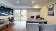 This single-floor leasing office is thoughtfully designed to provide a professional and welcoming environment for prospective residents. Rich hardwood flooring flows throughout, complementing a clean, modern aesthetic defined by recessed lighting and neutral tones accented with bold navy blue walls.

The main reception and workspace area serves as the heart of the office, featuring a large, contemporary L-shaped desk finished in a woodgrain laminate, paired with ergonomic seating for staff and comfortable upholstered guest chairs. Community floor plans and lifestyle imagery displayed on the walls offer visitors an immediate sense of the broader community. Large sliding glass doors flood the space with natural light and open to an exterior balcony, offering views of the surrounding neighborhood.

A dedicated seating lounge near the entrance features two leather accent chairs positioned alongside a bold decorative wall panel, creating an inviting waiting area. A pass-through window connects the main area to a secondary back room, enhancing communication across the office.

A separate conference or consultation room offers a glass-top table surrounded by tufted blue chairs beneath an elegant multi-shade chandelier, providing an intimate setting for one-on-one meetings. A small service area with white cabinetry and a marble-style countertop rounds out the space, offering practical support for daily operations.