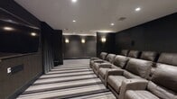 Located at 45 Creekside Ln in Malvern, PA, this meticulously designed private home theater offers an immersive cinematic experience within a refined, upscale setting. The space is accessed through a dramatic entrance corridor lined with dark textured walls, bold striped carpeting in tones of gray, taupe, and black, and pairs of ornate gold quatrefoil wall sconces that cast a warm, inviting glow. A decorative console table with a diamond-patterned base and a framed mirror anchor the entryway, setting an Art Deco-inspired tone carried throughout the space. The theater room itself continues the dark, moody aesthetic with deep charcoal wall paneling and floor-to-ceiling drapes framing the large wall-mounted display screen. Recessed ceiling lighting supplements the elegant wall sconces, providing layered illumination ideal for a theater environment. Multiple rows of plush, oversized leather power recliner seats with built-in cup holders are arranged to optimize sightlines to the screen, comfortably accommodating a generous number of viewers. A subwoofer and surround sound speakers complete the audio-visual setup, ensuring a fully immersive experience. The consistent use of rich materials, refined lighting, and thoughtful design details make this home theater a standout entertainment space.