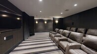 Located at 45 Creekside Ln in Malvern, PA, this meticulously designed private home theater offers an immersive cinematic experience within a refined, upscale setting. The space is accessed through a dramatic entrance corridor lined with dark textured walls, bold striped carpeting in tones of gray, taupe, and black, and pairs of ornate gold quatrefoil wall sconces that cast a warm, inviting glow. A decorative console table with a diamond-patterned base and a framed mirror anchor the entryway, setting an Art Deco-inspired tone carried throughout the space. The theater room itself continues the dark, moody aesthetic with deep charcoal wall paneling and floor-to-ceiling drapes framing the large wall-mounted display screen. Recessed ceiling lighting supplements the elegant wall sconces, providing layered illumination ideal for a theater environment. Multiple rows of plush, oversized leather power recliner seats with built-in cup holders are arranged to optimize sightlines to the screen, comfortably accommodating a generous number of viewers. A subwoofer and surround sound speakers complete the audio-visual setup, ensuring a fully immersive experience. The consistent use of rich materials, refined lighting, and thoughtful design details make this home theater a standout entertainment space.
