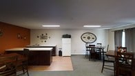 This single-floor space offers an open and functional layout that seamlessly blends a kitchen area with a communal dining space. The kitchen is equipped with dark wood cabinetry, a light-colored countertop with an integrated sink, and a full-size white refrigerator, providing all the essentials for meal preparation and everyday use. The flooring transitions from warm hardwood in the kitchen area to plush carpeting in the dining zone, clearly defining each functional space within the open floor plan. The dining area accommodates multiple guests with a sturdy dark wood dining table and matching cross-back chairs, making it well-suited for group gatherings. Recessed rectangular fluorescent light fixtures are mounted flush with the ceiling, ensuring bright, even illumination throughout the space. The neutral-toned walls are accented with decorative touches, including a striking oval metal wall medallion and a small arrangement of wall art, adding character and warmth to the overall aesthetic. The space presents a clean, uncluttered environment that balances practicality with a welcoming ambiance, making it an ideal setting for communal use.
