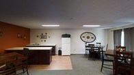 This single-floor space offers an open and functional layout that seamlessly blends a kitchen area with a communal dining space. The kitchen is equipped with dark wood cabinetry, a light-colored countertop with an integrated sink, and a full-size white refrigerator, providing all the essentials for meal preparation and everyday use. The flooring transitions from warm hardwood in the kitchen area to plush carpeting in the dining zone, clearly defining each functional space within the open floor plan. The dining area accommodates multiple guests with a sturdy dark wood dining table and matching cross-back chairs, making it well-suited for group gatherings. Recessed rectangular fluorescent light fixtures are mounted flush with the ceiling, ensuring bright, even illumination throughout the space. The neutral-toned walls are accented with decorative touches, including a striking oval metal wall medallion and a small arrangement of wall art, adding character and warmth to the overall aesthetic. The space presents a clean, uncluttered environment that balances practicality with a welcoming ambiance, making it an ideal setting for communal use.