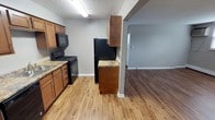 This single-floor space offers a practical, open-concept layout where the kitchen flows seamlessly into the living area, creating a connected and functional environment. The kitchen is equipped with medium-toned wood cabinetry providing ample storage, complemented by granite-look countertops that add a touch of character. Black appliances, including a gas range with overhead microwave and a full-sized refrigerator, are neatly integrated into the design. A textured ceiling adds dimension overhead, while light laminate flooring runs through the kitchen, transitioning to a deeper, darker laminate in the adjacent living area — a subtle design detail that visually distinguishes the two spaces. Neutral gray walls throughout create a clean, modern backdrop that pairs well with the warm cabinetry tones. The open sightline between the kitchen and living room enhances the sense of space, making the layout feel expansive and airy. White baseboards provide a crisp contrast against the flooring, finishing the space with a polished look. Overall, the unit presents a well-maintained, move-in ready environment with a cohesive and inviting aesthetic.