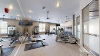 Located at 805 Canal St in Chattanooga, TN, this single-floor fitness center offers a thoughtfully designed exercise space with a well-rounded selection of equipment and a clean, modern aesthetic. The room features light-toned carpeting, neutral gray walls, and recessed lighting complemented by ceiling fans that help maintain a comfortable workout environment.

Cardio equipment is prominently featured throughout, including multiple treadmills, elliptical trainers, stair climbers, and Peloton spin bikes, giving users a wide variety of aerobic training options. A dedicated free weights area includes a full rack of dumbbells and a workout bench positioned on protective floor mats. Wall-mounted televisions and a refreshment vending unit add to the convenience of the space.

Large windows along one wall invite natural light and provide views of the surrounding neighborhood. A striking black-and-white photographic mural of the Chattanooga riverfront and bridge serves as a focal point, adding a strong sense of local character and visual interest. Full-length mirrors expand the sense of space and allow users to monitor their form during exercise. A separate enclosed area with frosted glass panels offers additional privacy for focused training. The overall atmosphere is clean, functional, and inviting, making it a well-appointed fitness facility.