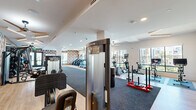 Located at 3421 N Freeway Blvd in Sacramento, CA, this generously sized fitness center offers a well-appointed, single-floor exercise facility designed to support a comprehensive workout experience. The space is thoughtfully divided into distinct zones, each tailored to specific fitness activities.

The cardio area features a row of treadmills, elliptical machines, stationary bikes with interactive screens, rowing machines, and a Xebex air runner, all positioned along large windows that flood the space with natural light and provide views of the surrounding palm-lined outdoor area. Ceiling fans enhance ventilation and comfort throughout.

The strength training zone is equipped with adjustable benches, a full rack of dumbbells reflected by floor-to-ceiling mirrors, cable machines, and functional training equipment including sled push tracks. A bold, geometric-patterned accent wall in warm earth tones adds a striking visual element to this area, complemented by light wood-look flooring and plush carpet underfoot.

A dedicated free-weight and stretching area features hardwood-style flooring, open space for flexibility work, and ample room for floor exercises. A built-in refreshment station with cabinetry and a mini-refrigerator provides added convenience. Modern linear lighting, recessed fixtures, and ceiling fans complete the clean, contemporary aesthetic of this well-equipped fitness facility.