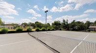 This outdoor recreational facility centers around a well-maintained hard-surface tennis court, clearly marked with regulation court lines and fitted with a standard net. The court surface is finished in a classic gray tone, providing clear visibility and a professional playing experience. A tall court light pole rises at the center baseline, ensuring the space can be utilized during evening hours. The court is fully enclosed by a chain-link perimeter fence, offering both safety and containment during play. Surrounding the court, mature deciduous trees and lush greenery create a natural, park-like setting that provides shade and a pleasant visual backdrop. The open-sky exposure allows for ample natural light throughout the day, complemented by the well-placed overhead lighting for after-hours use. In the background, a playground structure is visible, suggesting this tennis court is part of a broader community recreational complex. The combination of quality court infrastructure, thoughtful lighting, and verdant surroundings makes this an inviting and functional outdoor sports amenity suited for players of all skill levels.