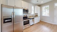 This two-story Tybee floor plan in Myrtle Beach, South Carolina offers a well-organized layout across two levels, designed to maximize functionality in a compact footprint. The main level features a kitchen equipped with white shaker-style cabinetry extending floor to ceiling, granite countertops, a stainless steel appliance package including a refrigerator, range, dishwasher, and over-the-range microwave, and a single-basin sink positioned beneath a window that brings in natural light. Light wood-tone flooring runs throughout the main living areas, creating a clean, cohesive look. An additional room on the main level features ceiling fan lighting and light wood flooring, offering flexible use as a living or dining space. Upstairs, the two bedrooms feature soft carpet, vaulted ceilings, ceiling fans, and windows that allow natural light. Each bedroom includes a closet with built-in shelving for organized storage. Both bathrooms are finished with granite-topped white vanities and tub-shower combinations with white surrounds. A staircase with a classic baluster railing connects the two levels. The home also includes an attached garage, adding convenient covered parking and extra storage space.