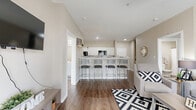 This single-floor unit offers five bedrooms and four bathrooms, efficiently designed to maximize functionality and comfort. The open-concept living area flows seamlessly into the kitchen, which features white cabinetry, a subway tile backsplash, white countertops, and black appliances including a gas range and built-in microwave. A spacious breakfast bar with seating for four serves as a natural divider between the kitchen and living space, making it ideal for both casual dining and socializing. The living room benefits from natural light through large windows and a glass-paneled entry door, creating a bright and welcoming atmosphere. Wood-style flooring runs throughout the unit, providing a cohesive and easy-to-maintain surface. Each bedroom is compact yet functional, with windows that bring in natural light, and several feature dedicated closets with built-in shelving for organized storage. The four bathrooms are consistently finished with white vanities, wood-style flooring, and shower-tub combinations. Additional conveniences include a dedicated laundry area and two hallways that efficiently connect all rooms. The neutral color palette of warm beige and white tones throughout lends the space a clean, modern aesthetic.