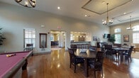 This single-story community clubhouse is thoughtfully designed to accommodate a variety of social and recreational activities. Throughout the space, rich hardwood flooring, soaring ceilings, and neutral gray-toned walls create a cohesive and welcoming atmosphere, while recessed lighting and stylish pendant and chandelier fixtures provide warm, well-distributed illumination.

The lounge area is anchored by tall arched windows that draw in abundant natural light, complemented by a wall-mounted flat-screen television, a large sectional sofa, patterned accent chairs, and a central coffee table. Rustic sliding barn doors connect zones seamlessly, adding architectural character.

The game room houses a full-size billiards table beneath a rustic chandelier, alongside dining tables and chairs that offer flexible seating for casual gatherings. A dedicated wall-mounted cue rack keeps the space organized and functional.

The dining area provides additional seating with dark wood tables and upholstered chairs, flowing naturally toward a pass-through bar counter with a granite surface and woven bar stools beneath pendant lighting—ideal for serving refreshments. Adjacent functional spaces are visible beyond, completing this well-appointed, multipurpose clubhouse.