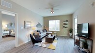 This single-floor space offers a clean, contemporary design with a warm and inviting ambiance throughout. The living area serves as the heart of the home, showcasing smooth wood-look flooring that runs seamlessly underfoot, complemented by neutral-toned walls that create a calm, versatile backdrop. A ceiling fan with an integrated light fixture anchors the room from above, providing both comfort and illumination. Natural light pours in through a large window fitted with horizontal blinds, brightening the space and creating an airy, open atmosphere. The room offers generous space for furnishings, as demonstrated by the comfortable sectional sofa, accent chair, and a central coffee table arranged atop a stylish geometric area rug. Wall-mounted shelving on one side of the room provides functional display space, while the overall layout encourages easy flow and movement. The white baseboards and clean wall finishes add a polished, well-maintained character to the interior. With its modern aesthetic, practical features, and thoughtfully designed layout, this single-floor space balances style and functionality to create a comfortable and welcoming environment.