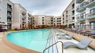 This outdoor amenity space is centered around a generously sized resort-style swimming pool with striking blue tile accents and crystal-clear water. The pool is framed by a wide, open sundeck outfitted with rows of modern grey lounge chairs and side tables, offering ample space for relaxation. Multiple entry points with stainless steel railings provide easy pool access throughout the deck area.

Surrounding the pool, the courtyard extends into several distinct seating zones. Shaded dining areas feature wicker-style tables and chairs beneath vibrant red umbrellas, ideal for outdoor gatherings. A dedicated lounge lawn area is furnished with plush sectional sofas and accent chairs accented with bold red cushions, set on a manicured artificial grass surface beneath a large canopy umbrella — creating a comfortable, living-room-like outdoor retreat.

Mature shrubs, ornamental trees, and landscaped garden beds with iron fencing line the perimeter, adding a sense of privacy and greenery throughout the courtyard. Classic lamp posts are positioned throughout the space, ensuring the area is well-lit in the evenings.

The entire outdoor area is enclosed within a U-shaped, multi-story building featuring contemporary architecture with a combination of brick and smooth facade finishes, private balconies, and large windows — contributing to an intimate yet open courtyard atmosphere.