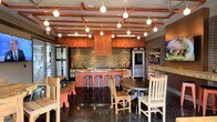 This single-floor space showcases a bold industrial-rustic aesthetic that seamlessly blends raw materials with warm, earthy tones. The ceiling is a standout architectural feature, where exposed copper pipes run in organized horizontal lines across the full span of the room, complemented by an array of globe pendant lights that cast a warm, ambient glow throughout the space.

At the heart of the room sits a central kitchen island finished in a rich terracotta-red hue, fitted with a row of metal bar stools that invite casual seating. The island is framed by a dramatic textured backsplash in stacked stone, while a matching copper-toned range hood serves as a focal centerpiece above. Full-sized stainless steel appliances flank the cooking area, adding a modern contrast to the rustic surroundings.

The flooring is a polished dark concrete, reflecting light and enhancing the sense of depth within the space. Dining areas are furnished with a mix of wooden tables, benches, and chairs in natural and painted finishes, reinforcing the eclectic, handcrafted character. An exposed brick wall to the right adds additional texture, while large windows allow natural light to filter in, brightening the overall atmosphere.
