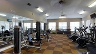 This fitness center offers a comprehensive workout environment designed to accommodate a range of exercise needs. The space features vaulted ceilings that contribute to an open, airy feel, while two ceiling fans provide ventilation throughout. Multiple windows along the walls allow natural light to fill the room, creating a bright and energizing atmosphere.

The flooring consists of a checkered pattern of two-tone carpet tiles in tan and brown, which helps define the workout area while providing cushioning underfoot. A large wall-to-wall mirror along one side visually expands the space and allows users to monitor their form during exercise.

The equipment selection is well-rounded, including a multi-station cable and weight machine for strength training, an adjustable bench press, and a dedicated free weights section with a full dumbbell rack. A stability ball and a cardio machine, likely a stationary bike or rowing machine, round out the available equipment. Fluorescent lighting supplements the natural light to ensure consistent illumination throughout the facility, making this a functional and inviting space for daily fitness routines.