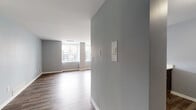 This single-floor residence presents a clean, contemporary aesthetic defined by cool gray walls, white trim, and a thoughtful mix of flooring throughout. The open living area features wood-style plank flooring and is anchored by a ceiling fan with integrated lighting. A peninsula kitchen opens directly to the living space, offering a light quartz countertop with a built-in sink, dark shaker-style cabinetry, and stainless steel appliances including a refrigerator and range. The layout promotes a natural flow between the kitchen and living areas.

The bedrooms are finished with plush carpet and benefit from large windows that frame urban city views, filling each room with natural light. One bedroom includes a wall-mounted panel and additional built-in features. The bathroom is well-appointed with a wide dark-toned vanity topped with a white quartz surface, a large mirror with a multi-bulb light bar overhead, a soaking tub with tile surround, and wood-style flooring that ties into the rest of the unit. An intercom panel near the entryway adds a practical functional element. Overall, the space maintains a consistent modern palette with durable finishes and urban views throughout.