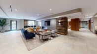 This expansive single-floor amenity space is designed with a modern aesthetic, blending warm wood accents, stone tile flooring, and bold contemporary furnishings throughout. The open-plan layout flows seamlessly between distinct functional zones, each thoughtfully curated for both socializing and relaxation.

The central lounge area anchors the space with a sleek, wall-mounted linear fireplace set against a concrete surround, flanked by a striking reclaimed wood accent wall. Comfortable seating — including a deep navy sofa, tan swivel chairs, and crisp white accent chairs — invites gathering and conversation. A wall-mounted flat-screen television completes the entertainment setup.

Adjacent to the lounge, a generously sized bar area features a curved, mosaic-clad island with a white quartz countertop and bold yellow barstools. Warm wood wall paneling, a mounted television, and stainless steel appliances give this area a polished yet inviting feel. A separate curved reception-style counter with additional yellow bar seating and pendant lighting adds to the dynamic layout.

The game room offers a full-size billiard table beneath an elegant geometric chandelier, accompanied by a built-in banquette seating nook and reclaimed wood columns with integrated sconce lighting.

A more intimate meeting or dining room features rich dark wall finishes, a round table with yellow upholstered chairs, lush greenery, and a warm wood built-in shelving unit — ideal for private gatherings or focused work sessions.