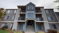 This single-floor residence is situated within a well-maintained multi-story building, distinguished by its blue and gray lap siding exterior, private balconies, and a central stairwell entry. The building's façade showcases a gabled roofline accent and large windows that lend the structure a clean, contemporary curb appeal.

Inside, the unit features a neutral color palette throughout, with warm wood-look flooring visible in the entryway area. Interior doors are fitted with sleek black lever-style hardware, contributing to a modern aesthetic that carries through the space.

Storage is a notable feature of this unit. The closet system is thoughtfully designed, incorporating multiple adjustable shelving units with clean white finishes alongside a sturdy metal hanging rod — providing well-organized, functional storage capacity. The closet space is generous, with both shelf and hanging sections making efficient use of the available area.

The entryway presents a tidy transitional zone, with crisp white walls, a clean-lined interior door, and wood-look flooring that sets a welcoming tone upon entry. The overall interior maintains a bright, minimalist atmosphere, achieved through consistent use of white and light gray tones across walls, trim, and built-in elements. The unit offers a practical and well-considered layout suited for comfortable everyday living.