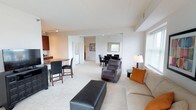 This single-floor residence offers a thoughtfully designed layout that balances comfort and functionality. The open-concept living and dining area is bright and welcoming, with soft carpet flooring, neutral walls, and an abundance of natural light streaming through large windows and a sliding glass door that opens to an outdoor balcony with greenery views. The living room provides generous space for seating and entertainment, centered around a clean, contemporary arrangement.

The kitchen is well-equipped with rich dark cherry wood cabinetry extending to the ceiling for ample storage, granite-toned countertops, stainless steel appliances including a full-size refrigerator, a double-basin sink, dishwasher, and track lighting overhead. A breakfast bar adds a casual dining option alongside the separate dining area.

The bedrooms are carpeted and serene, each featuring windows with outdoor views, closet space, and room for full bedroom furniture sets. The primary bedroom includes an en-suite bathroom with a long vanity, dark wood cabinetry, and a walk-in shower with sliding glass doors and tile flooring. A secondary half-bath with a single vanity and toilet serves the common areas. Throughout, the space maintains a consistent, neutral palette that creates a calm and inviting atmosphere.