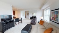 This single-floor residence offers a thoughtfully designed layout that balances comfort and functionality. The open-concept living and dining area is bright and welcoming, with soft carpet flooring, neutral walls, and an abundance of natural light streaming through large windows and a sliding glass door that opens to an outdoor balcony with greenery views. The living room provides generous space for seating and entertainment, centered around a clean, contemporary arrangement.

The kitchen is well-equipped with rich dark cherry wood cabinetry extending to the ceiling for ample storage, granite-toned countertops, stainless steel appliances including a full-size refrigerator, a double-basin sink, dishwasher, and track lighting overhead. A breakfast bar adds a casual dining option alongside the separate dining area.

The bedrooms are carpeted and serene, each featuring windows with outdoor views, closet space, and room for full bedroom furniture sets. The primary bedroom includes an en-suite bathroom with a long vanity, dark wood cabinetry, and a walk-in shower with sliding glass doors and tile flooring. A secondary half-bath with a single vanity and toilet serves the common areas. Throughout, the space maintains a consistent, neutral palette that creates a calm and inviting atmosphere.