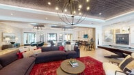 Located on Bonnar Bridge Parkway in Burlington, NC, this sophisticated single-floor clubhouse is designed to impress with a seamless blend of modern elegance and social functionality. The open-concept layout is anchored by a spacious lounge area featuring large, plush sectional sofas set atop a bold red area rug, complemented by striking orb-style chandeliers that hang from an ornate textured tray ceiling. Natural light filters through floor-to-ceiling sliding glass doors that open to an outdoor terrace, enhancing the airy atmosphere throughout.

A dedicated game room includes a full-size pool table surrounded by curated gallery wall art, with additional seating for relaxed socializing. The space flows naturally into multiple dining areas, including a formal dining zone with a large oval table, teal upholstered chairs, and gold-accented furnishings beneath another sculptural chandelier.

A modern kitchen bar area features clean white quartz countertops, warm wood cabinetry, and built-in beverage coolers, with a pass-through window connecting to an adjacent lounge. Textured stone accent walls run throughout, adding warmth and architectural depth. Conference-style seating and co-working surfaces round out the space, making it equally suited for productive gatherings and leisure.