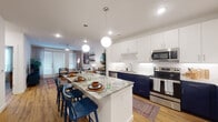 This well-appointed single-floor residence offers a thoughtfully designed open-concept layout that seamlessly connects the kitchen, dining, and living areas. The kitchen is a standout feature, showcasing a striking two-tone design with crisp white upper cabinetry and bold navy lower cabinets, complemented by a white subway tile backsplash and granite countertops. A generously sized central island with additional open shelving provides ample prep and dining space, illuminated by two globe pendant lights overhead. Stainless steel appliances, including a refrigerator, range, microwave, and dishwasher, complete the fully equipped kitchen. Light-toned hardwood flooring runs throughout the space, enhancing the bright and airy atmosphere created by recessed lighting and large windows. The living area offers generous natural light through floor-to-ceiling French doors and sizable windows fitted with blinds, and is anchored by a ceiling fan. The bedroom is a serene retreat featuring a large window, ceiling fan, and warm ambient lighting. The full bathroom continues the navy and granite design theme, pairing a soaking tub and shower combination with a well-lit vanity mirror. A spacious walk-in closet with multi-tiered shelving and hanging rods provides abundant storage.
