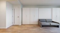 Located on the 30th floor of a high-rise in Seattle's First Hill neighborhood, this thoughtfully designed studio unit at 707 Terry Ave maximizes every square foot with a clean, modern aesthetic. Light hardwood flooring runs throughout, complementing the crisp white walls and recessed lighting that keep the space feeling bright and open.

The long, well-lit hallway leads into the main living area, where a built-in wall of floor-to-ceiling white cabinetry provides generous storage alongside an integrated Murphy bed and a compact gray sofa — a smart, space-efficient layout. The kitchen is outfitted with flat-front white cabinetry extending to the ceiling, stainless steel appliances including a full-size refrigerator, built-in oven, microwave, and an induction cooktop, all set against light stone countertops.

The bathroom features terrazzo-style tile flooring, a wall-mounted floating vanity with an illuminated mirror, and a full soaking tub with a handheld shower — finished in a clean, contemporary palette of white and soft grey. An in-unit laundry area and a private balcony offering city views round out this efficiently designed urban residence.