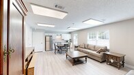 Located at 710 Lynn Dr SE in Jacksonville, Alabama, this single-floor space efficiently combines a kitchen and living area into one well-designed unit. Light-toned hardwood-style flooring runs throughout, creating a cohesive and clean aesthetic that ties the two functional zones together seamlessly.

The living area is bright and inviting, benefiting from a large window fitted with horizontal blinds that allows generous natural light to fill the room. A skylight overhead further enhances the brightness of the space. A textured ceiling adds subtle architectural character, while neutral beige walls provide a warm, understated backdrop.

The kitchen area is thoughtfully appointed with white shaker-style cabinetry — both upper and lower — featuring glass-front accent doors. Granite-look countertops provide ample prep space, complemented by a stainless steel double-basin sink and a stainless steel refrigerator. A microwave and dishwasher round out the functional appliances. A window above the counter brings in additional natural light, making the kitchen feel open and airy. A round dining table with cushioned chairs is positioned nearby, creating a practical dining area within the combined space.

An interior doorway connects to adjacent areas, and an exit door provides convenient access. Overall, the space presents a clean, functional, and welcoming environment with modern finishes and efficient use of its footprint.