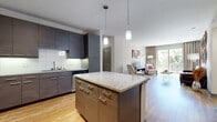This single-floor residence showcases a thoughtfully designed open-concept layout with contemporary finishes throughout. The kitchen serves as a central hub, anchored by a granite-topped island with pendant lighting overhead. Dark-toned, flat-front cabinetry lines the walls, complemented by a white tile backsplash and a full suite of stainless steel appliances, including a French door refrigerator, range, microwave, and dishwasher. Light hardwood flooring flows seamlessly from the kitchen into the living and dining areas, enhancing the sense of openness. The living room offers generous wall space and large windows dressed with floor-length drapes, allowing ample natural light. A dedicated dining nook accommodates a round table and chairs. The two bedrooms feature plush carpeting and large windows that invite natural light, providing a calm retreat. Both bathrooms are finished with granite-topped dark vanities, tile flooring, and tub-shower combinations surrounded by stone-look tile surrounds. One bathroom also includes a built-in closet with shelving. The overall design balances modern aesthetics with functional living spaces.