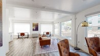 This thoughtfully designed single-floor space blends a professional and welcoming atmosphere through clean, modern finishes and a well-organized layout. The main open area serves dual purposes, functioning as both a comfortable lounge and a productive workspace. Warm cognac leather seating — including a full sofa and tufted accent chairs — is arranged over a vintage-style area rug, anchored by a minimalist hairpin-leg coffee table. Decorative wall art adds color and character to the otherwise crisp white walls. Multiple workstations with desks and ergonomic chairs line the perimeter, benefiting from generous natural light streaming through large windows that offer views of the surrounding streetscape. Window roller shades provide privacy and light control as needed. A sleek wall-mounted fireplace and a set of circular metallic wall accents add a stylish focal point to the lounge side of the room. The kitchenette features white shaker-style cabinetry with black hardware, dark countertops, a white subway tile backsplash, a stainless steel sink, and a compact refrigerator — ideal for light refreshments. The ADA-accessible restroom is clean and functional, equipped with grab bars and a wall-mounted soap dispenser. Wood-look luxury vinyl flooring runs throughout, tying all areas together with a warm, cohesive aesthetic.