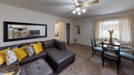 Located at 2196 Rockdell Dr. in Fairborn, Ohio, this single-floor unit offers a well-organized layout with a comfortable mix of living, dining, sleeping, and utility spaces. The living room serves as the central hub, featuring carpet flooring, a ceiling fan with light fixture, and an open sightline into the dining area. Natural light filters in through a window dressed with sheer curtains, creating a bright and welcoming atmosphere. The dining area accommodates a round glass table with seating for four, accompanied by a window with vertical blinds and sheer draping that allows additional natural light. The kitchen is outfitted with white cabinetry, a gas range, full-size refrigerator, a sink with a window above it, and warm hardwood-style flooring that adds character to the space. The bedrooms feature carpet flooring and are decorated in a consistent yellow and dark-toned palette, with windows providing natural light. One bedroom includes a painted cinder block accent wall, adding a distinctive architectural element. The full bathroom features hardwood-style flooring, a single-sink vanity with wood cabinetry, tiled walls, and a shower-tub combination. A utility area with an electrical panel and tiled walls completes the layout.