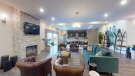 Located at 626 Lehman St in Columbus, Ohio, this single-floor community clubhouse offers a thoughtfully designed gathering space with a variety of amenities and contemporary finishes. The expansive main lounge serves as the heart of the space, anchored by a stacked stone fireplace with a wall-mounted TV, tufted leather armchairs, and a plush teal sectional sofa arranged over a large area rug. Recessed lighting, wall sconces, and a modern globe pendant chandelier provide warm, layered illumination throughout. Natural light pours in through full-height glass doors that offer views of the outdoor surroundings. Adjoining the lounge is a game area featuring a pool table and shuffleboard, set against a bold yellow accent wall with pendant lighting and an additional mounted TV, creating a lively entertainment zone. A corridor with wood-look flooring connects to additional rooms, accented by a large decorative clock and arched mirror. The fully equipped kitchen showcases deep navy shaker-style cabinetry, granite countertops, a subway tile backsplash, stainless steel appliances, an undermount sink, and globe pendant lights — combining function with refined style. Throughout the space, durable wood-look flooring, tray ceiling details, and curated décor contribute to a cohesive, upscale ambiance ideal for community gatherings.