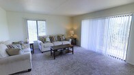 This single-floor residence offers a well-appointed living space designed for both comfort and functionality. The living room serves as the heart of the home, featuring wall-to-wall carpeting that adds warmth underfoot and contributes to the overall cozy ambiance. Large vertical-blind-covered windows flood the room with natural light, offering views of surrounding greenery, while a full-length sliding glass door provides convenient access to the outdoors and further enhances the sense of openness. The neutral color palette throughout — from the soft, light-toned walls to the understated flooring — creates a versatile backdrop that complements a variety of furnishing styles. The space is thoughtfully laid out to accommodate comfortable seating and a central coffee table arrangement, making it ideal for everyday living and casual entertaining. Ambient lighting from a table lamp adds a warm, inviting glow to the room during evening hours. The overall design emphasizes simplicity and livability, with clean lines and practical finishes throughout. The abundance of natural light, connection to the outdoors, and open layout combine to make this a bright and pleasant place to call home.
