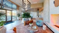 Located at 18750 Delaware St in Huntington Beach, California, this single-floor space serves as a sophisticated lobby and hallway that blends contemporary design with coastal warmth. The generously sized layout offers an open, airy atmosphere defined by soaring ceilings and expansive floor-to-ceiling windows that flood the interiors with natural light and provide sweeping views of the surrounding greenery.

The central lounge area anchors the space with a striking cylindrical metal chandelier descending from the high ceiling, encircling a lush, raised indoor planter brimming with tropical foliage. Curved seating in warm cognac leather and soft blue upholstered chairs are arranged around a marble-topped coffee table, creating an inviting social setting. A textured, multi-tonal area rug adds warmth to the polished tile flooring beneath.

A dedicated conference or meeting area features a tiled, stone-topped communal table surrounded by aqua-toned ergonomic chairs, accented by warm wood-paneled ceilings and a built-in display shelving unit with decorative metalwork. Adjacent to this is a corridor lined with natural wood cabinetry, a beverage station, and continued access to outdoor views through a full wall of windows. The overall ambiance is modern, welcoming, and thoughtfully curated with a refined coastal aesthetic.