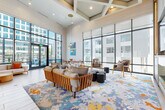 This expansive single-floor amenity lounge is designed with a contemporary aesthetic that balances energy and comfort. The architecture is immediately distinguished by dramatically tall ceilings accented with coffered detailing and shiplap wall paneling in crisp white, creating a clean, airy backdrop throughout the space. Floor-to-ceiling glass walls and oversized windows flood the interior with abundant natural light while offering unobstructed views of the surrounding urban environment.

A striking sculptural installation of multicolored geometric shapes cascades across the soaring wall above the bar area, serving as a bold focal point. The bar itself features a sleek stone-topped island with warm yellow bar stools, dark cabinetry, and under-counter refrigeration, making it well-suited for social gatherings. Adjacent to the bar is a dedicated dining area furnished with a solid wood table, woven-seat chairs in warm tones, and woven pendant lighting overhead, complemented by vibrant aquatic artwork on the walls.

The lounge zones feature a large, colorful abstract area rug anchoring a collection of mid-century-inspired wood-framed sofas, lounge chairs, and organic wood stump accent tables. A built-in media wall with open shelving and an integrated flat-screen television provides an entertainment focal point. The warm wood-look flooring ties all zones together, creating a cohesive and inviting communal space.