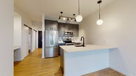 This single-floor space showcases a thoughtfully designed modern interior with clean lines and a cohesive contemporary aesthetic. The open-concept kitchen serves as a focal point, featuring charcoal gray flat-front cabinetry with matte black hardware, full stainless steel appliances including a French door refrigerator, electric range, and over-the-range microwave. A geometric patterned tile backsplash adds visual interest behind the cooking area, while a large white waterfall-edge island with an undermount sink and matte black faucet provides additional workspace and a natural gathering point. Globe and track pendant lighting illuminates the kitchen with a modern touch.

Light wood-tone flooring runs continuously throughout the space, contributing to a warm, cohesive feel. The bedroom is bright and airy, benefiting from a large picture window that frames urban views and floods the room with natural light. A built-in closet with shelving provides ample storage. The bathroom is finished with a white quartz countertop, an undermount sink, gray vanity cabinetry with matte black hardware, and a wide frameless mirror. The overall layout, visible in the floorplan scan, is compact yet efficiently organized, offering a well-connected flow between living areas.
