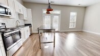 This contemporary apartment offers a blend of style and functionality. The kitchen is a standout feature, boasting crisp white cabinetry, subway tile backsplash, and high-end stainless steel appliances including a full-size refrigerator, range, and built-in microwave. The space is illuminated by trendy pendant lighting, creating an inviting atmosphere for cooking and dining.

The bathroom continues the modern aesthetic with a marble-look tiled shower, glass partition, and a vanity with a dark countertop contrasting against white cabinetry. Wood-look flooring extends throughout the apartment, providing warmth and durability.

The living area is bathed in natural light thanks to large windows, some extending from floor to ceiling. High ceilings and neutral wall colors contribute to a sense of spaciousness. The room's minimalist design allows for flexible furnishing options.

A unique sliding barn door adds a touch of industrial chic to the interior. The apartment's open concept layout promotes a seamless flow between living spaces, ideal for modern lifestyles. Overall, this unit combines contemporary design with practical amenities in a bright, airy setting.