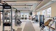 This thoughtfully designed fitness center offers a comprehensive workout environment spread across a single, expansive floor. The space is organized into distinct training zones, each tailored to a specific type of exercise. The cardio area lines the perimeter walls, where large windows flood the room with natural light and provide outdoor views, creating an energizing atmosphere for workouts. This section is outfitted with a full lineup of treadmills, ellipticals, stationary bikes, and stair climbers, complemented by wall-mounted flat-screen televisions for entertainment during training sessions. The strength and functional training area occupies the central portion of the floor, anchored by a large TKO multi-station power cage complete with a heavy bag, kettlebells, and medicine balls. Additional cable machines, weight benches, and a barbell rack with plates provide a full range of resistance training options. The flooring transitions between rubberized sport surfaces and smooth fitness flooring depending on the zone. Architecturally, the space features a distinctive tiered tray ceiling with integrated linear LED lighting that adds depth and a modern, polished aesthetic. Soaring ceiling heights in select areas further enhance the sense of openness. The overall ambiance is clean, bright, and professional, making it a well-appointed facility suited for a wide variety of fitness routines.