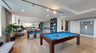 Located on the 22nd floor in Baltimore, MD, this generously proportioned lounge offers an expansive, open-plan layout across a single level, designed to accommodate a variety of social and recreational activities. The space is finished with wide-plank light hardwood flooring throughout, creating a warm, cohesive aesthetic that complements the modern interior design.

The lounge is divided into several distinct functional zones. A dedicated seating area features plush sofas and accent chairs arranged around a glass coffee table, anchored by a large area rug in gold tones. Tall windows along this section flood the space with natural light and offer elevated views of the Baltimore skyline. A built-in fireplace with large-format tile surround and flanking open bookshelves with bold teal geometric-patterned backing serves as a central architectural focal point.

The game area houses a full-size pool table and additional gaming options, lit by track lighting above. A raised platform section with industrial-style bistro tables and stools adds a dynamic, multi-level element to the layout, accented by a striking deep-blue patterned feature wall with curated artwork.

A wrap-around wet bar with a herringbone wood-paneled base, white countertop, and wall-mounted television offers a sophisticated hospitality station with ample bar seating. Recessed lighting throughout maintains a bright, welcoming ambiance across every zone of this well-appointed lounge.