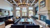 Located at 1155 Museum Blvd in Vernon Hills, IL, this single-floor community clubhouse delivers an upscale, well-appointed amenity space defined by thoughtful design and high-end finishes. The centerpiece of the space is a grand lounge area boasting soaring ceilings accented by a striking coffered and reclaimed wood ceiling detail, anchored by a large circular chandelier. Tall arched windows flood the room with natural light and offer views of the outdoor patio, while a classic fireplace with an ornate mantel adds warmth and elegance. Rich hardwood flooring, tufted leather sofas, and deep navy velvet seating create a refined yet welcoming atmosphere. A media lounge nook features wall-mounted TV, banquette seating, and gold-accented wall sconces for an intimate viewing experience. The business center offers a large collaborative table, individual computer workstations, built-in shelving with cozy booth seating, and globe pendant lighting, all bathed in natural light from arched windows. A sleek wet bar area showcases bold graphic wallpaper, dark cabinetry with marble-look countertops, and stainless steel appliances. The open-plan layout is unified by black-framed glass partition doors, open-display shelving, and a cohesive palette of navy, gold, and neutral tones throughout.