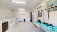 Located at 1335 Water Tower Circle in Greenville, South Carolina, this professional pet grooming studio is thoughtfully designed to accommodate all aspects of pet care in a single, well-appointed space. The room features a smooth, easy-clean floor finished in a light blue-grey tone, complemented by neutral-colored walls and bright fluorescent lighting that ensures excellent visibility throughout. A standout feature is the dedicated bathing alcove, which includes a built-in walk-in pet wash basin with a hand-held sprayer, flanked by tile-clad walls that extend partway up for durability and easy maintenance. Adjacent to the bathing area, a stainless-steel grooming table with a paw-print mat provides a functional and sanitary work surface, equipped with grooming arm attachments and blow-dry tools. A wall-mounted mini-split HVAC unit ensures year-round climate control. The studio is accessible through both a set of glass double doors facing a green outdoor view and an interior glass sliding door connecting to an adjacent room. Whimsical dog-themed artwork adorns the walls, adding a welcoming touch. The space is efficiently organized to support a full grooming workflow, making it ideal for professional pet grooming services.