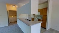 This single-story space centers around a practical, open-concept kitchen designed for everyday functionality. The kitchen features a prominent peninsula with a neutral laminate countertop and a double-basin sink, serving as both a workspace and a natural divider between the kitchen and adjacent living areas. A structural column anchors the peninsula, adding architectural definition to the layout.

The cooking area is equipped with white appliances, including an electric range with overhead microwave, a refrigerator, and ample cabinetry finished in warm honey-toned wood. The cabinetry provides generous storage along the perimeter walls, complementing the efficient use of space throughout the kitchen.

Flooring transitions from tile in the kitchen area to carpet in the surrounding spaces, creating distinct zones within the open floor plan. A dedicated laundry area is visible in the background, housing a full-size washer and dryer — a notable convenience within the unit itself.

Lighting is provided through flush-mount ceiling fixtures, maintaining a clean and straightforward aesthetic throughout. The overall design is functional and well-organized, prioritizing practicality and ease of use.