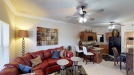 This single-floor residence offers an open-concept design that integrates the living area and kitchen into one cohesive, functional space. The living room is a welcoming focal point, anchored by a rich red leather sectional sofa accented with decorative pillows in warm earth tones. A pair of bold black-and-white graphic side tables adds a striking contrast against a patterned area rug, creating a curated, stylish atmosphere. A large abstract canvas in warm gold and brown tones adorns the wall, adding visual depth and character. Crown molding runs throughout the space, lending an elegant, finished look to the interior, while a ceiling fan with an integrated light fixture ensures comfort and practical illumination. The kitchen flows naturally from the living area and features warm honey-toned wood cabinetry, a dark countertop breakfast bar with seating, and black appliances including a built-in microwave. The bar seating, consisting of white upholstered chairs, provides a casual dining option that bridges the kitchen and living space. Natural light filters into the interior, enhancing the warm, inviting ambiance. Overall, this space offers a well-appointed and thoughtfully designed single-level layout suited for comfortable everyday living.