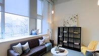 This single-floor space makes a strong impression with its notably high ceilings and tall, expansive windows that flood the living area with abundant natural light and offer views of surrounding greenery. The architectural design blends modern and industrial elements, evident in the exposed ceiling details and clean wall finishes.

The living area is thoughtfully arranged, anchored by a dark tufted sofa accented with decorative throw pillows and a cozy blanket, conveying a relaxed yet styled atmosphere. A sleek, round white coffee table sits at the center, providing functional surface space without overwhelming the room. Beside the sofa, a large window ledge doubles as a casual seating or display area, further maximizing the use of space.

A substantial dark grid-style shelving unit lines one wall, offering generous open storage and display space for books and decorative objects. A large artistic map-style canvas hangs above it, adding a graphic, urban-inspired focal point. A slender pendant light descends from the high ceiling, contributing warm ambient lighting. The overall ambiance is contemporary, calm, and well-curated.
