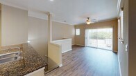 This single-floor residence offers a thoughtfully designed layout that balances open communal spaces with comfortable private areas. The living room is a welcoming centerpiece, featuring warm hardwood-style flooring, crown molding, and a ceiling fan. A sliding glass door with vertical blinds opens to an outdoor balcony, filling the space with natural light and providing views of the surrounding greenery. A secondary window adds additional brightness to the room.

The kitchen is efficiently designed with rich cherry wood cabinetry, dark granite countertops, a tile backsplash, and black appliances including a range and refrigerator. Track lighting illuminates the workspace, while a double stainless steel sink completes the functional layout. A peninsula with granite countertop connects the kitchen to the living and dining area, enhanced by decorative columns that frame the open-concept flow.

The bedrooms are carpeted for comfort, with warm neutral tones carried throughout. The primary bedroom benefits from a walk-in closet and a generously sized window with outdoor views. Additional bedrooms offer ample space and natural light through windows fitted with blinds.

The bathroom is finished with tile flooring, a tiled tub-shower combination, a rich wood vanity with granite countertop, and a framed mirror, providing a clean and functional space. Crown molding is a recurring detail that lends a polished touch throughout the home.