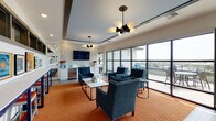 Located at 11001 W 15th Pl in Lakewood, Colorado, this sophisticated single-floor community lounge offers a thoughtfully designed gathering space that blends modern aesthetics with comfort. The main lounge area is anchored by a bold, vibrant orange area rug that contrasts elegantly with the teal velvet upholstered chairs and sofas arranged around a marble-topped coffee table, creating an inviting and stylish seating environment. A striking textured accent wall serves as a focal point, framing a wall-mounted flat-screen television above a wet bar with white cabinetry, a sink, and a stainless steel refrigerator — ideal for entertaining. Distinctive sculptural chandelier lighting adds a modern, artistic touch to the tray ceiling overhead. Along one wall, a high bar counter with bar stools sits beneath a curated gallery of framed artwork, providing a casual perch for socializing. Floor-to-ceiling glass panel walls and doors flood the interior with abundant natural light and offer unobstructed views of the surrounding Lakewood skyline. These doors open directly onto a furnished outdoor terrace, seamlessly extending the living space and offering an al fresco retreat. Light wood-tone flooring anchors the transitional areas, completing the polished, contemporary ambiance of this inviting communal space.